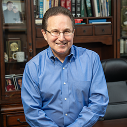 Dr. Kenneth P. Martinez poses seated on his desk in front of a bookcase with photos and books