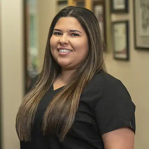 A young woman with long light brown hair wears black short-sleeved scrubs and smiles at the camera