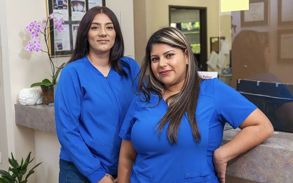 2 women pose in blue scrubs. They lean against the front desk.