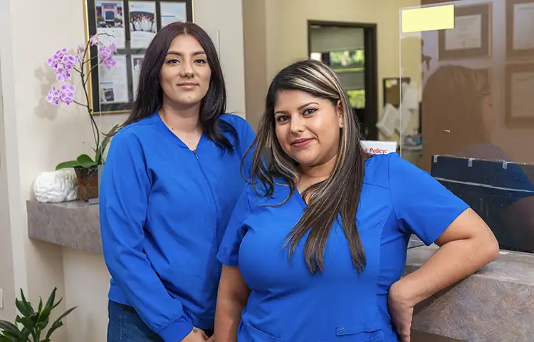 2 women pose in blue scrubs. They lean against the front desk.