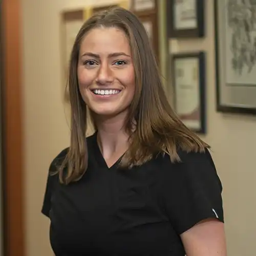 A young woman with long light brown hair wears short-sleeved black scrubs and smiles at the camera