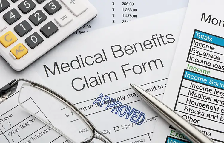 Close-up of medical benefits claim forms, a calculator, pen, and pair of reading glasses.