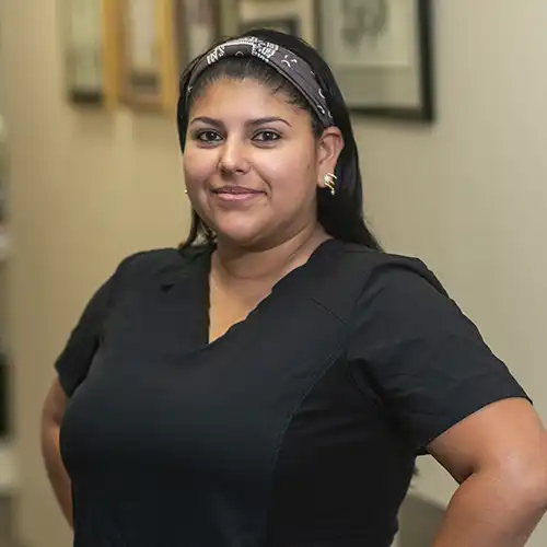 A young woman with long dark hair tied up with a bandana headband smiles at the camera. She wears short-sleeved black scrubs