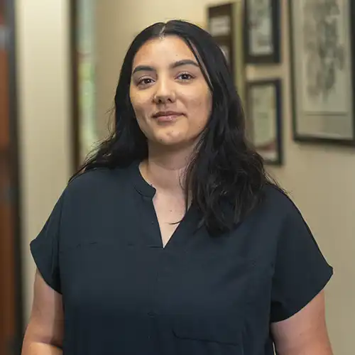 A young woman with long dark hair wearing short-sleeved black scrubs smiles shyly at teh camera
