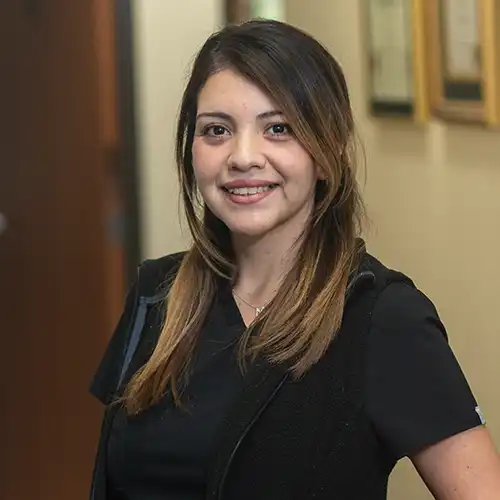 A young woman with long light brown hair wears short-sleeved black scrubs and smiles at the camera
