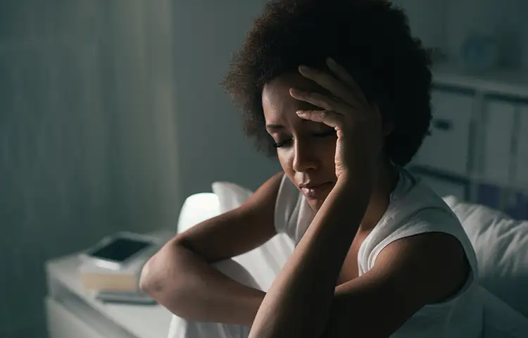 Black woman sitting in a darkened room with a pained expression and one hand to her forehead.