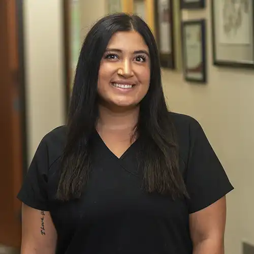 A young woman with long dark brown hair wears short-sleeved black scrubs and smiles at the camera