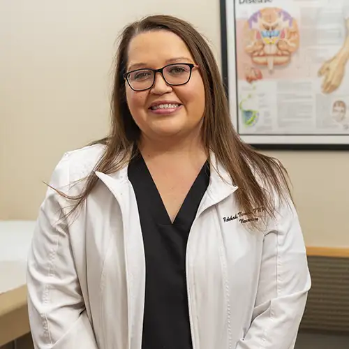 Rebekah Torres stands in her exam room wearing a white lab coat. An illustrated informational poster showing the brain is on the wall behind her.