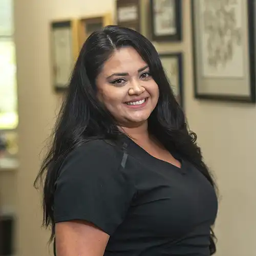 A woman with long dark hair wearing short-sleeved black scrubs stands three-quarters to the camera and smiles over her shoulder