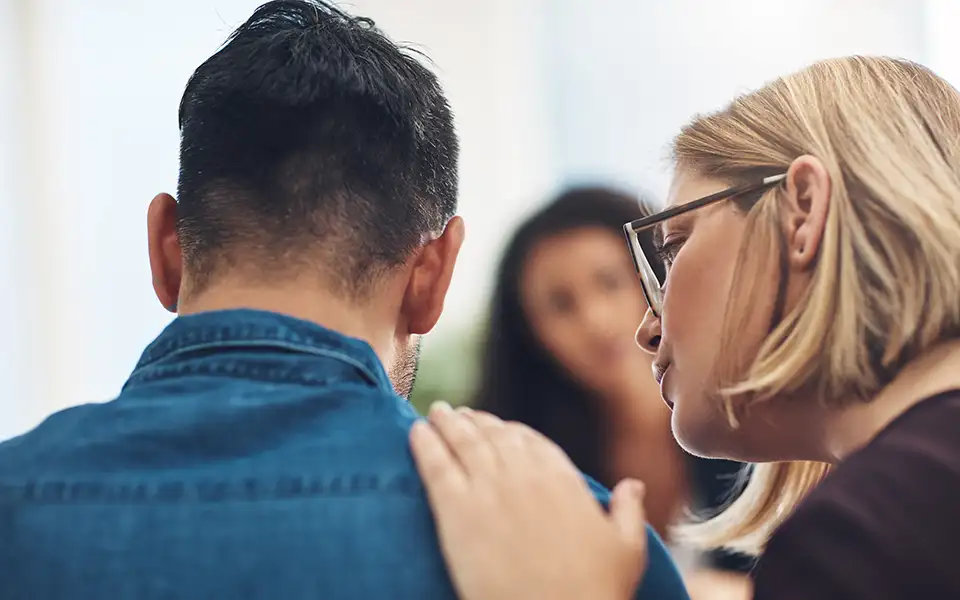 A man with short dark hair in a chambray shirt is seen from the back. A woman with blonde chin-length hair leans over and puts her hand on his shoulder.