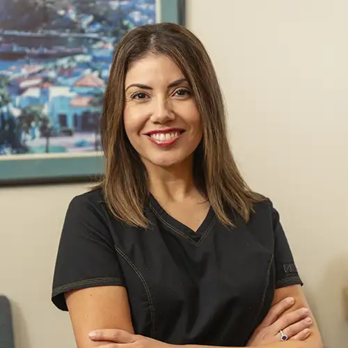 Vanessa Madrigal poses in her exam room with her arms folded. She wears black, short-sleeved scrubs and a painting is behind her on the wall.