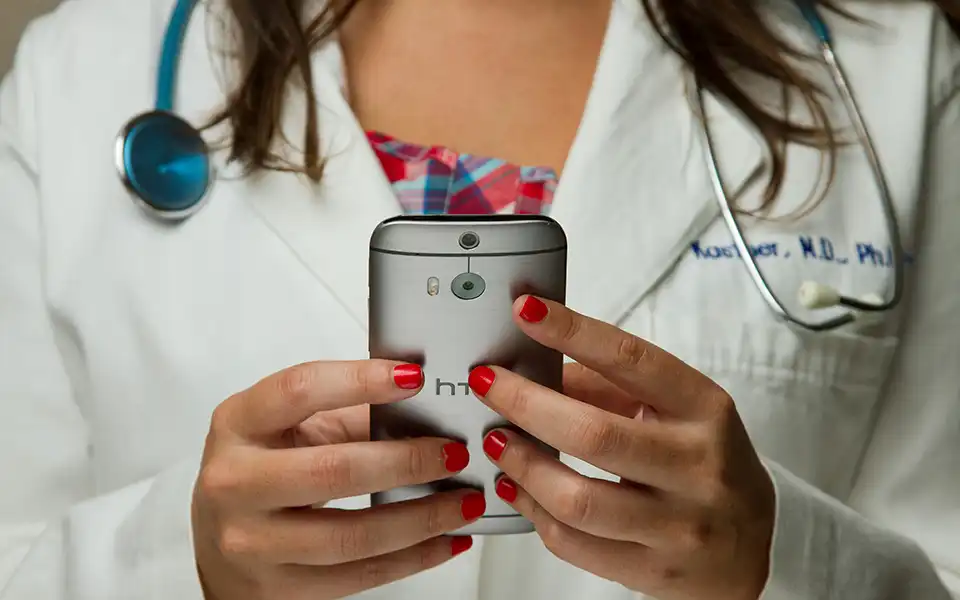 Tight shot of a female doctor's hands holding a mobile phone in front of her to speak to a patient over video. The doctor wears a white lab coat and has a stethoscope around her neck.