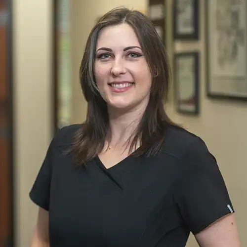 A woman with shoulder-length medium brown hair wearing black short-sleeved scrubs smiles at the camera