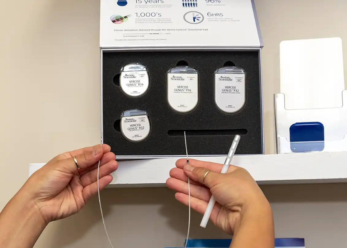 Hand holding a sample wire for DBS devices. A display case of Boston Scientific DBS devices is on the shelf behind the hands.