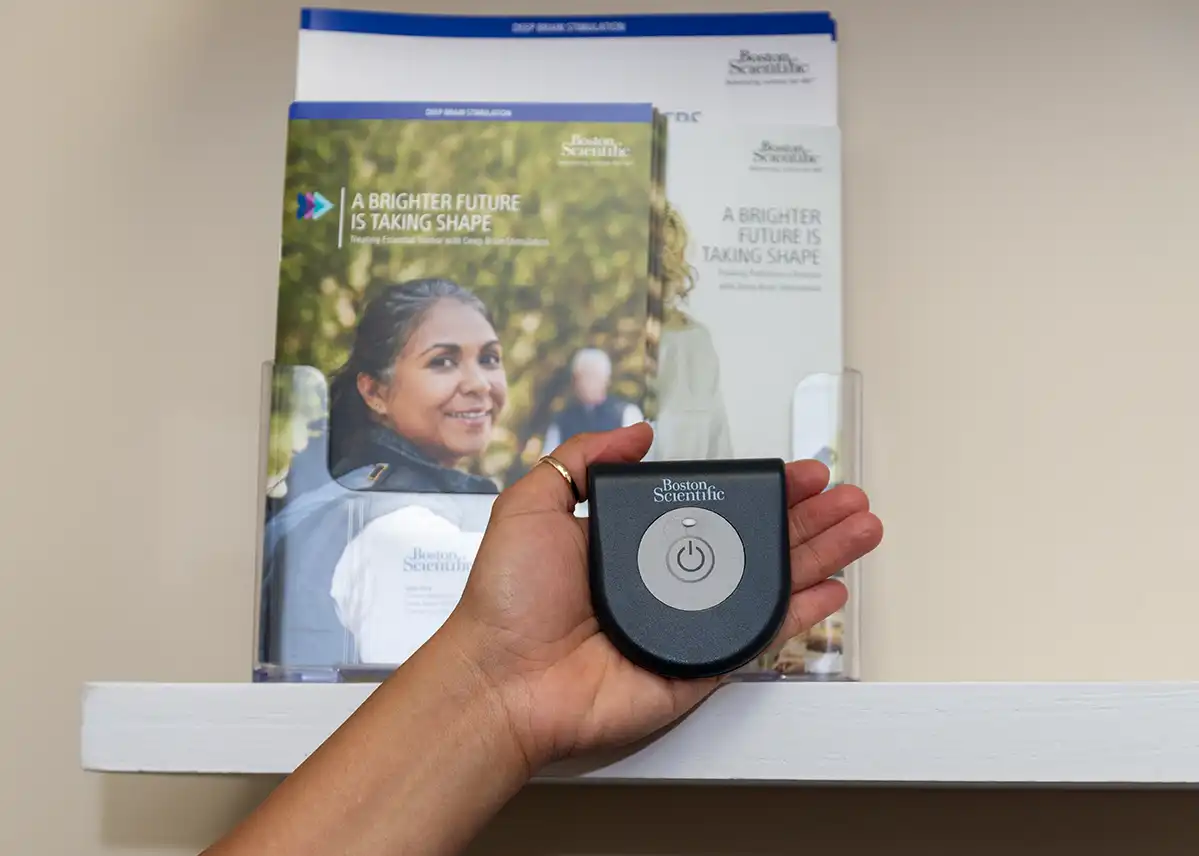 A hand holding up a palm-sized dark grey medical device with a light grey circle in the center. It says Boston Scientific and a brochure about the device is on the shelf behind the hand.