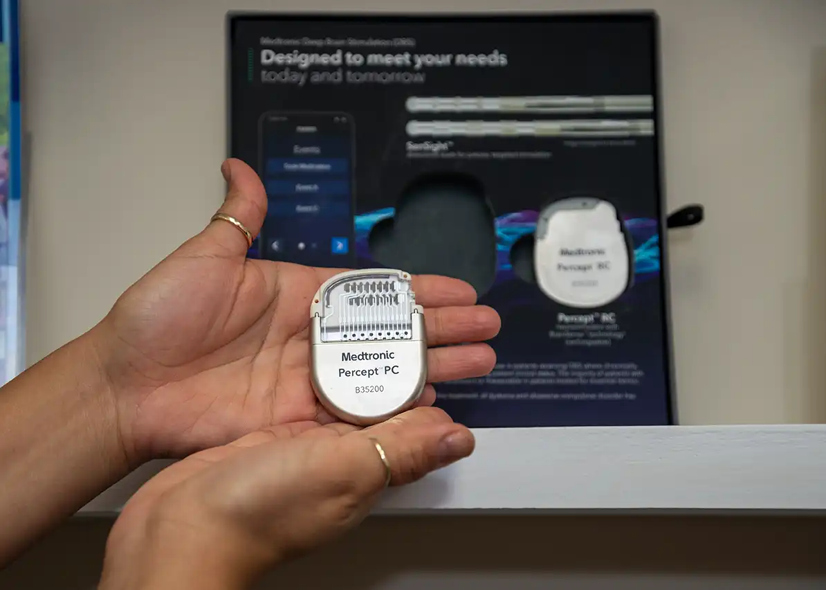 Two hands hold a small silver medical device about 2 inches tall and 1.5 inches wide. A brochure is on a shelf behind the hands.