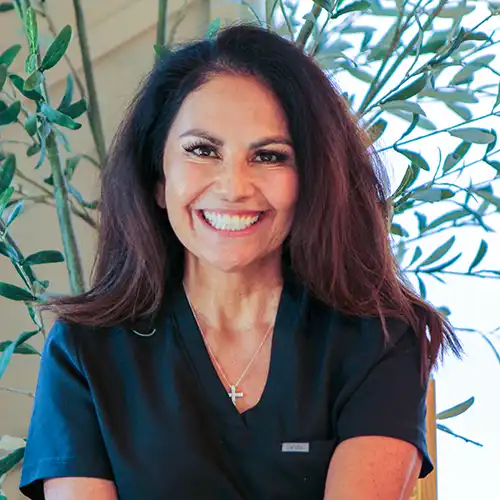 A middle-aged woman with shoulder-length brown hair wears short-sleeved black scrubs and smiles at the camera. Behind her is a bamboo plant.