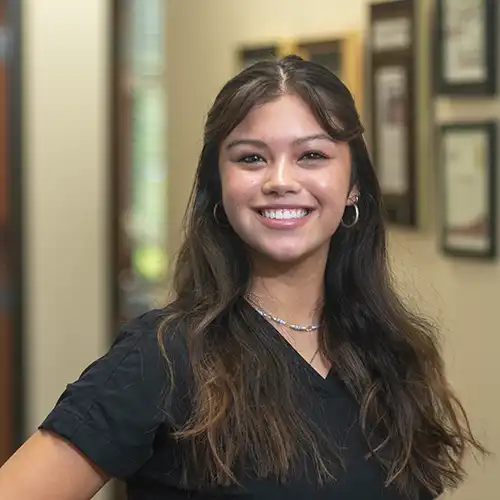 A young woman with long brown hair wears a short-sleeved set of scrubs and smiles at the camera
