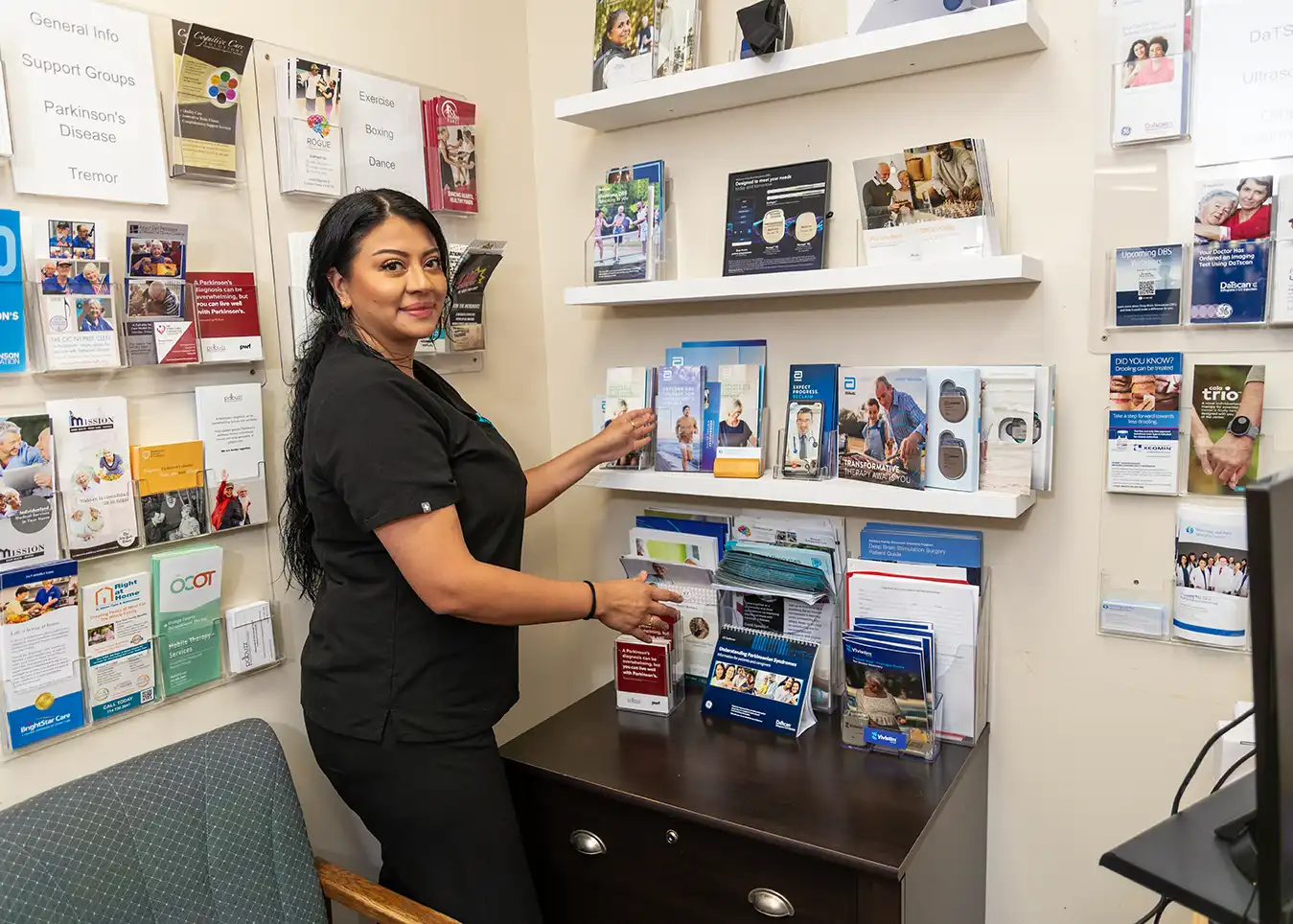 A woman with long dark hair stands in a corder with medication brochures displayed over both walls.