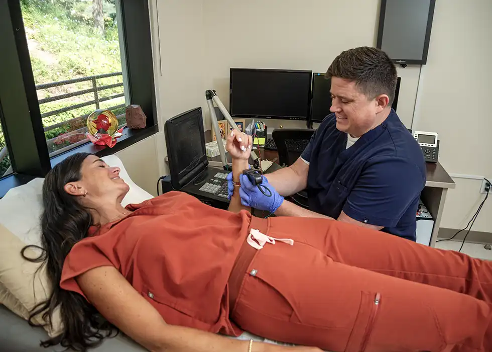 A woman with long, dark hair lays down on an examination table while a neurodiagnostic technician sits next to the exam table in front of a computer. He holds a medical device against her wrist.