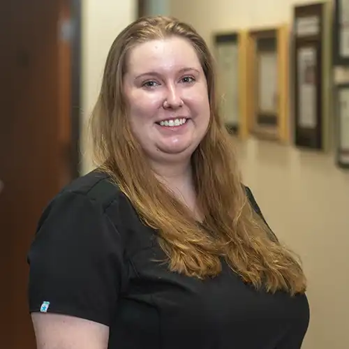 A young woman with long red hair looks over her shoulder and smiles at the camera. She wears short-sleeved black scrubs.